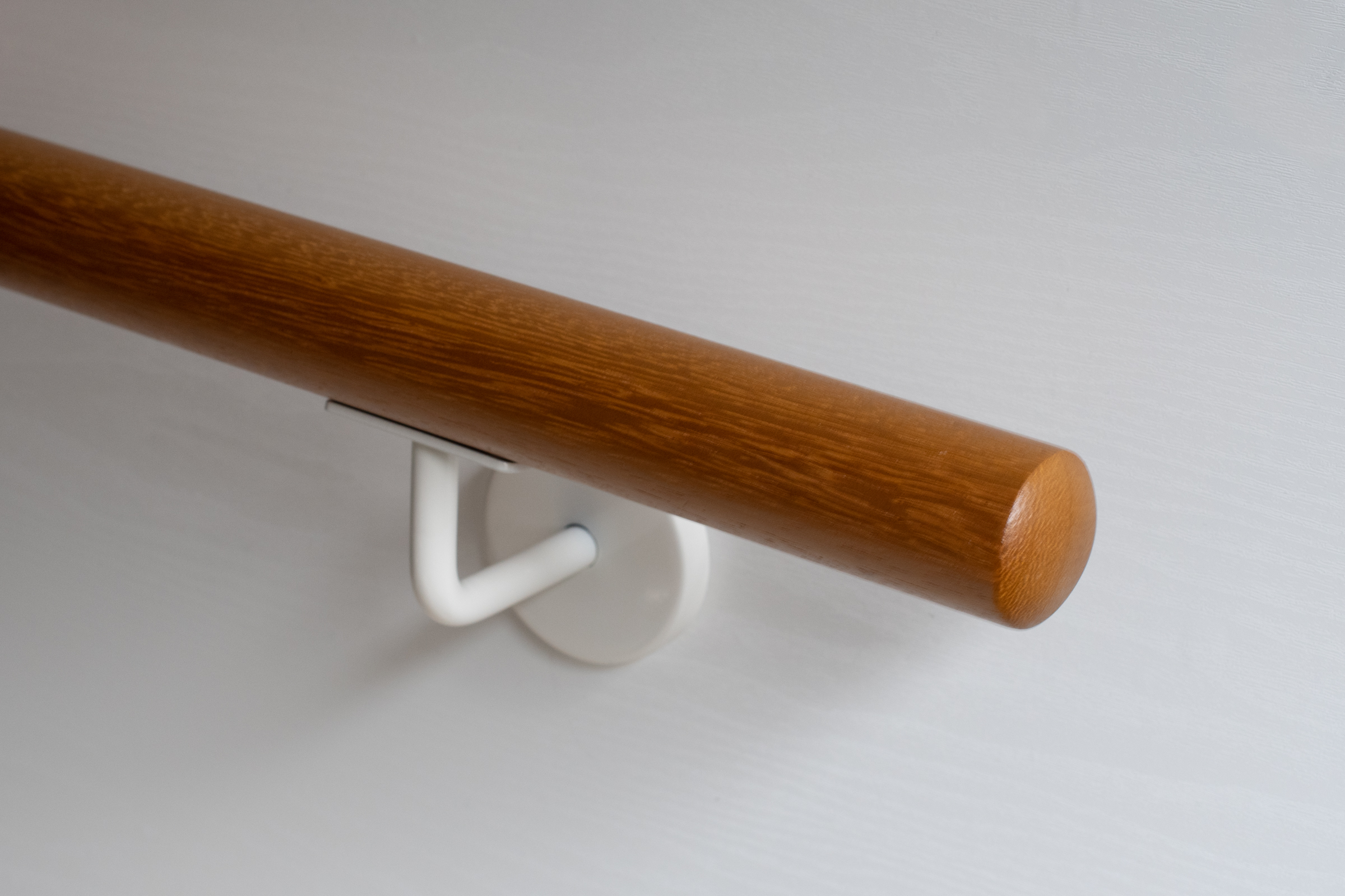 Handlauf Kambala rund mit weiß lackiertem Wandhalter an weißer Wand, goldbraunes Holz, Nahaufnahme des Endes