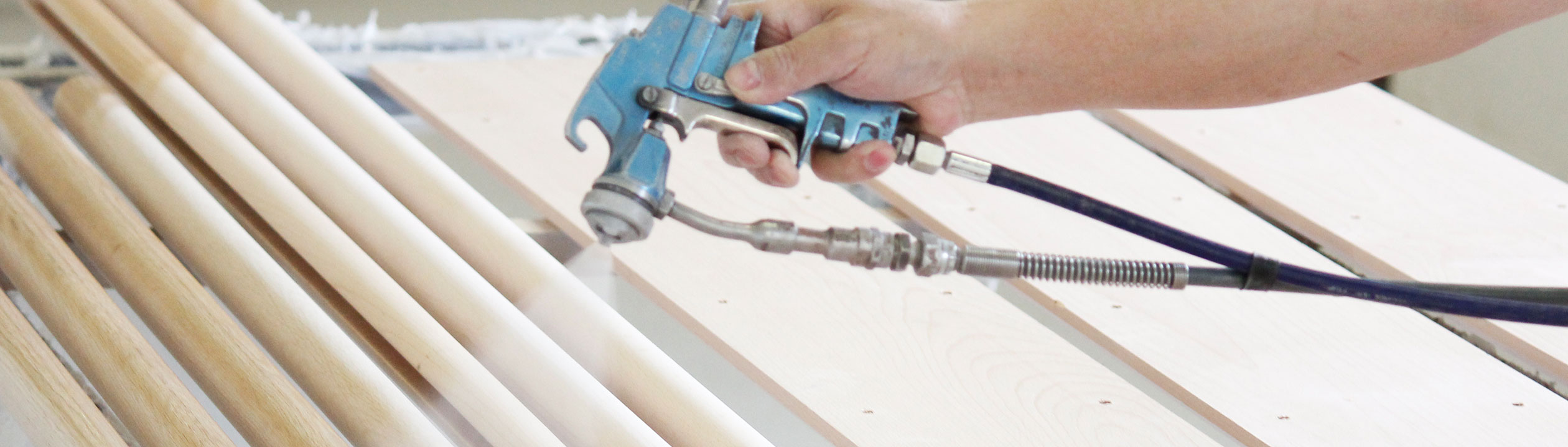 Handlaufproduktion: Holzhandläufe werden mit der Spritzpistole lackiert, Hand mit Lackierpistole über hellen Holzstäben
