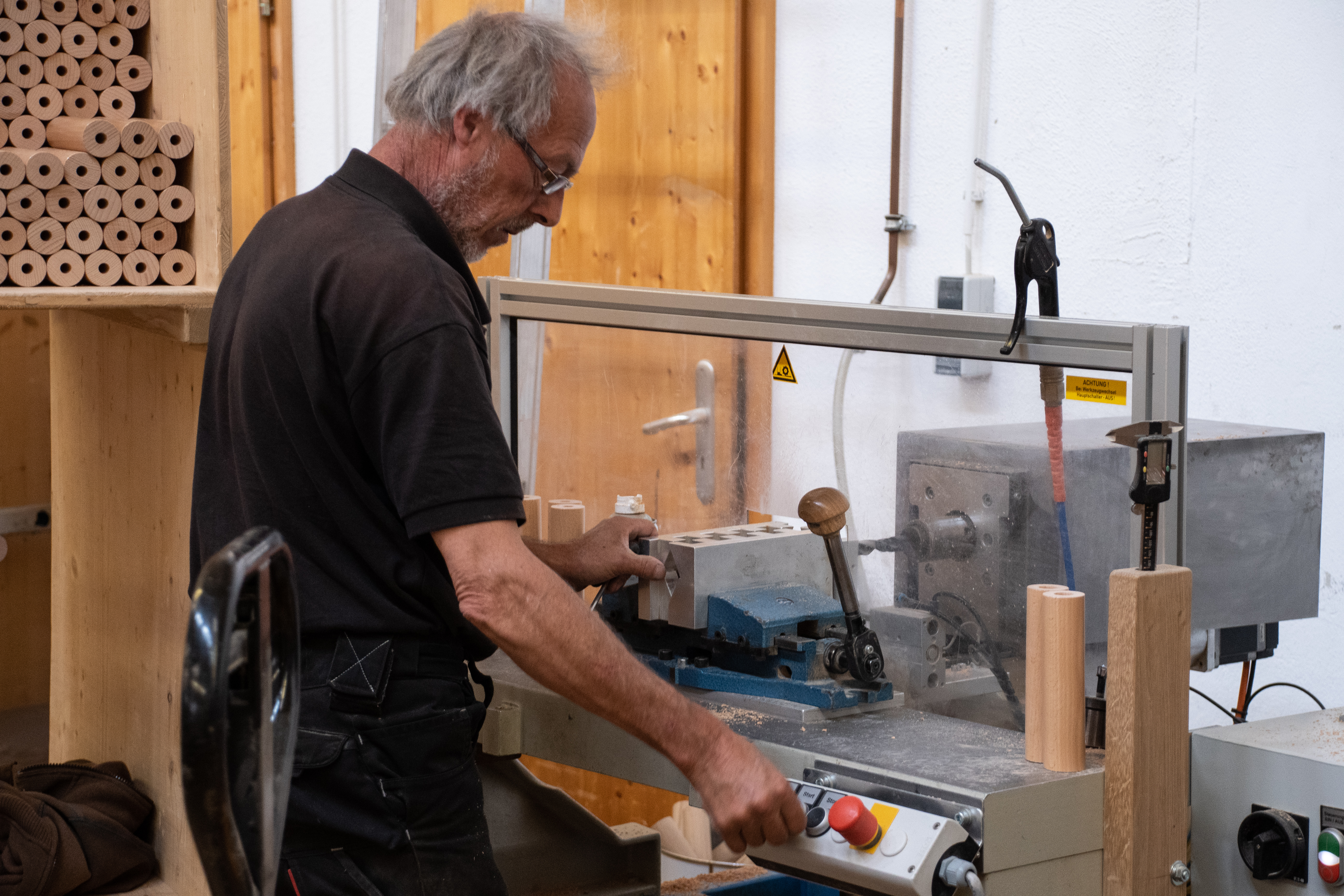 Handlaufherstellung: Mitarbeiter bohrt ein Holzteil an der Bohrmaschine in der Werkstatt, Holzregal mit Rundhölzern hinten