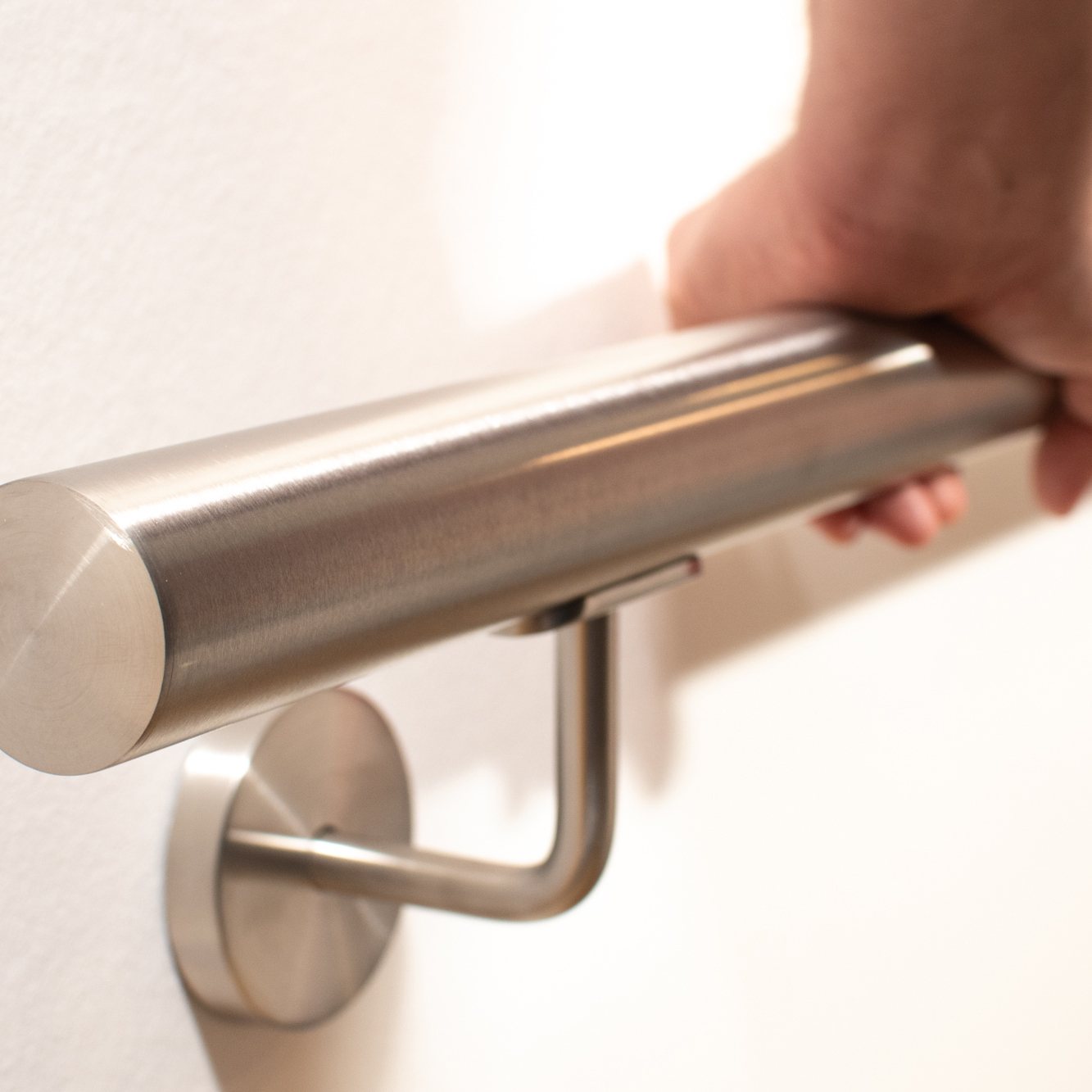 Handlauf Edelstahl rund an weißer Wand mit Edelstahl-Wandhalter, Hand greift den gebürsteten Rohrhandlauf