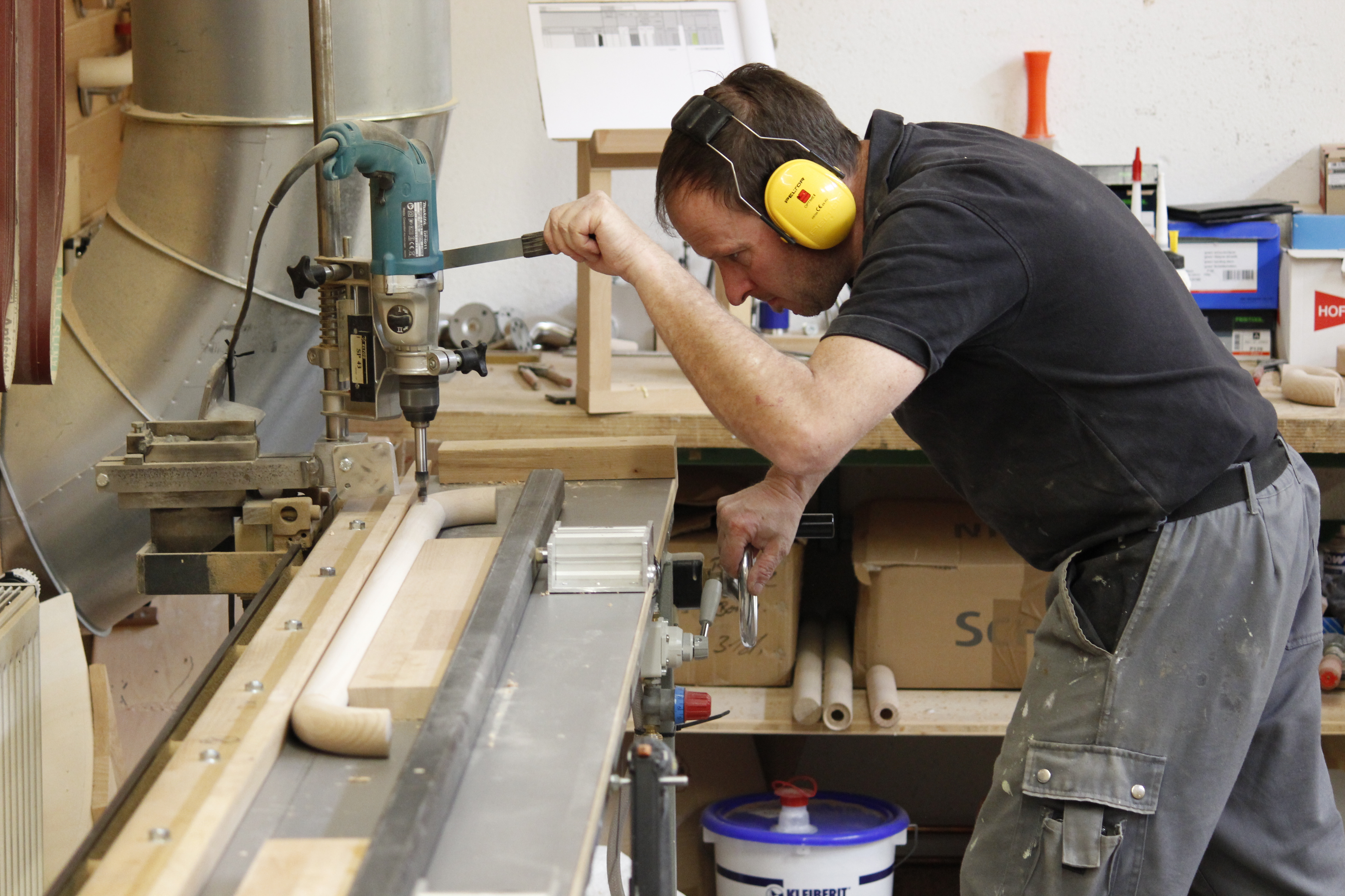 Handwerker mit Gehörschutz bohrt in der Werkstatt an einer Ständerbohrmaschine Löcher in einen Holzhandlauf